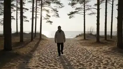 A man in his sixties walking through pines to a calm Baltic shore in the morning and stopping to look at the water